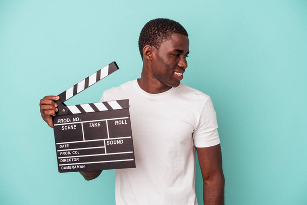 Młody Afroamerykanin trzymający klapperboard na niebieskim tle wygląda na uśmiechniętego, wesołego i miłego. - Zdjęcie, obraz