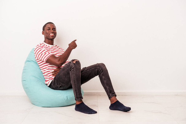 Young African American man sitting on a puff isolated on white background smiling and pointing aside, showing something at blank space. - Foto, Imagem