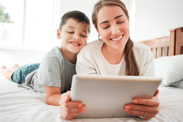 Happy caucasian mother and son holding digital tablet while lying together on a bed. Little boy and mom watching movie online or playing game while spending time at home. - Zdjęcie, obraz
