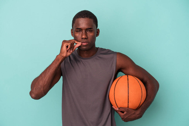 Young African American man playing basketball isolated on blue background with fingers on lips keeping a secret. - 写真・画像