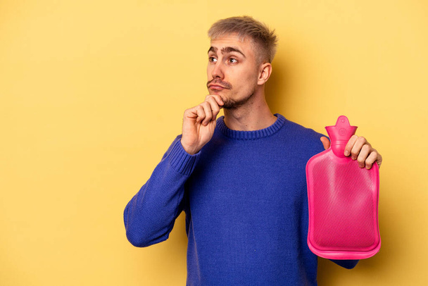 Young caucasian man holding a water bag isolated on yellow background looking sideways with doubtful and skeptical expression. - Foto, afbeelding