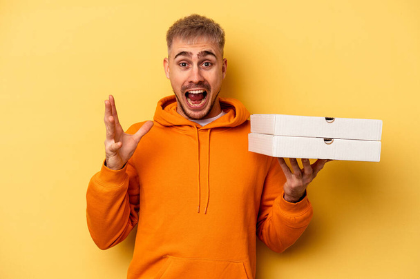 Young caucasian man holding pizzas isolated on yellow background receiving a pleasant surprise, excited and raising hands. - Fotó, kép