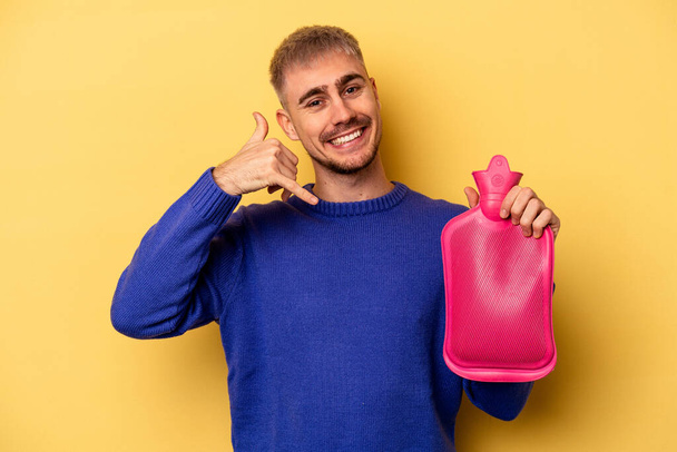 Young caucasian man holding a water bag isolated on yellow background showing a mobile phone call gesture with fingers. - Fotografie, Obrázek