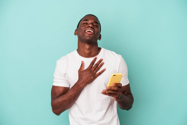 Jonge Afro-Amerikaanse man met een mobiele telefoon geïsoleerd op blauwe achtergrond lacht luid houden hand op de borst. - Foto, afbeelding