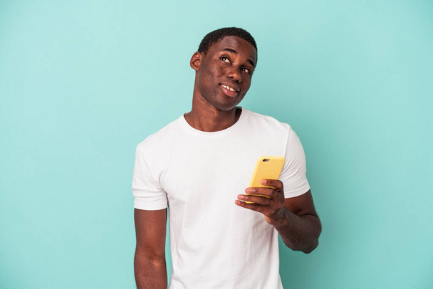 Νεαρός Αφροαμερικανός που κρατά ένα κινητό τηλέφωνο απομονωμένο σε μπλε φόντο ονειρεύεται την επίτευξη στόχων και σκοπών - Φωτογραφία, εικόνα
