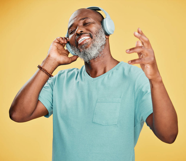 Szczęśliwy, dojrzały Afroamerykanin noszący słuchawki i słuchający muzyki tańczący na żółtym tle w studiu. Uśmiechnięty czarny człowiek czuje się wolny podczas wyrażania poprzez taniec. - Zdjęcie, obraz