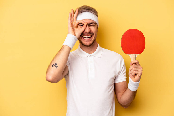 Young caucasian man holding ping pong rackets isolated on yellow background excited keeping ok gesture on eye. - Foto, afbeelding