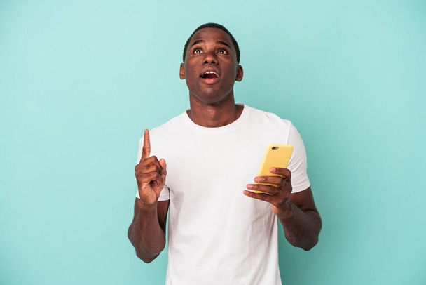 Giovane uomo afroamericano in possesso di un telefono cellulare isolato su sfondo blu che punta verso l'alto con la bocca aperta. - Foto, immagini