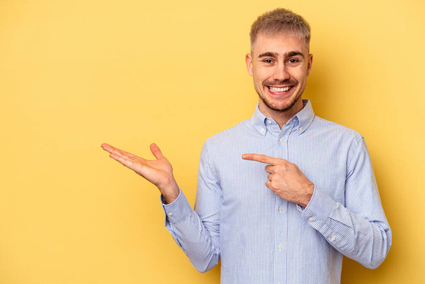 Young caucasian man isolated on yellow background excited holding a copy space on palm. - Foto, immagini