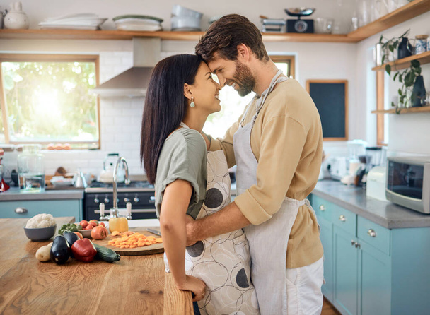 Happy interracial couple touching foreheads while sharing romantic intimate moment at home while wearing aprons and cooking together. Young caucasian man and hispanic wife looking into each others - Photo, Image