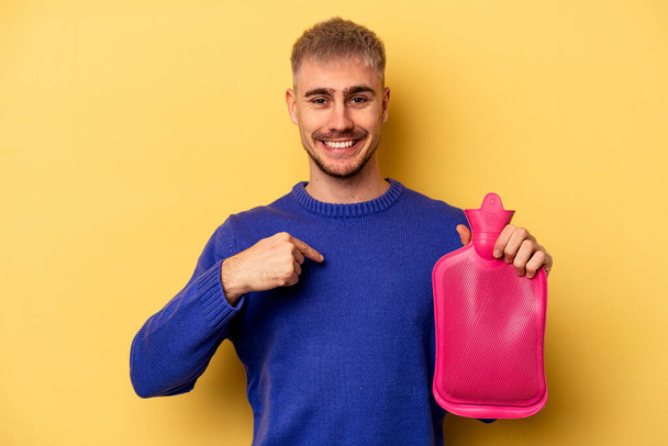 Young caucasian man holding a water bag isolated on yellow background person pointing by hand to a shirt copy space, proud and confident - 写真・画像