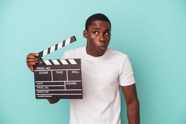 Young African American man holding clapperboard isolated on blue background shrugs shoulders and open eyes confused. - Photo, Image