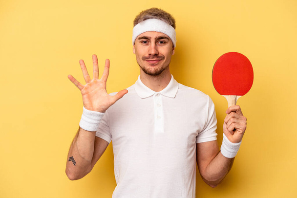 Fiatal kaukázusi férfi kezében ping-pong ütők elszigetelt sárga háttér mosolygós vidám bemutató száma öt ujjal. - Fotó, kép