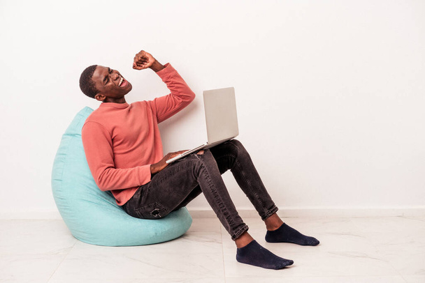 Junger Afroamerikaner sitzt mit Laptop auf weißem Hintergrund auf einem Puff und hebt nach einem Sieg die Faust, Siegerkonzept. - Foto, Bild