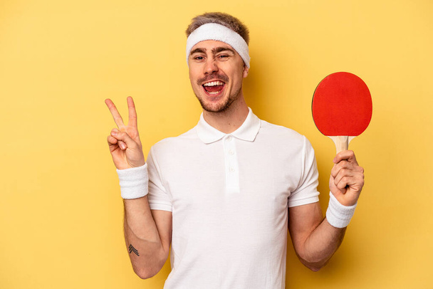 Junger kaukasischer Mann hält Tischtennisschläger isoliert auf gelbem Hintergrund und zeigt fröhlich und unbeschwert ein Friedenssymbol mit Fingern. - Foto, Bild