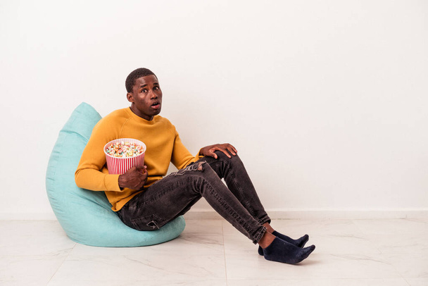 Mladý Afroameričan sedí na obláčku jíst popcorn izolované na bílém pozadí pokrčí rameny a otevřené oči zmatené. - Fotografie, Obrázek