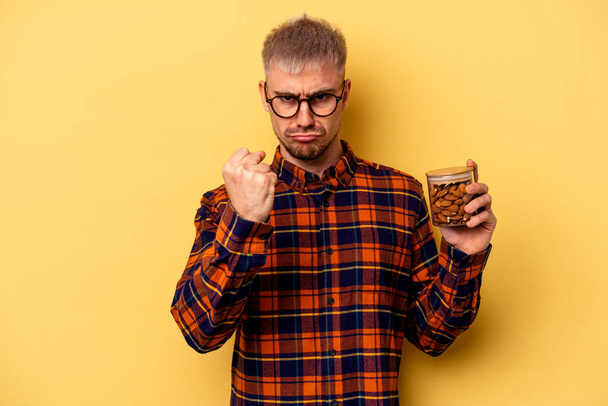 Young caucasian man holding a almond jar isolated on yellow background showing fist to camera, aggressive facial expression. - Photo, Image