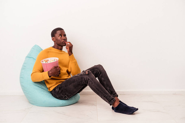 Молодой афроамериканец, сидящий на слоеном попкорне на белом фоне, расслабленно думает о чем-то, глядя на пространство для копирования.. - Фото, изображение