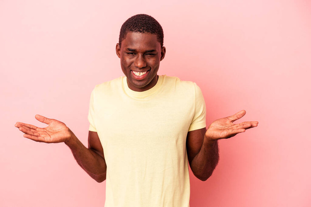 Joven afroamericano aislado sobre fondo rosa hace escala con los brazos, se siente feliz y confiado. - Foto, Imagen