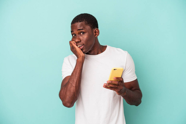 Jonge Afro-Amerikaanse man met een mobiele telefoon geïsoleerd op blauwe achtergrond bijten nagels, nerveus en zeer angstig. - Foto, afbeelding