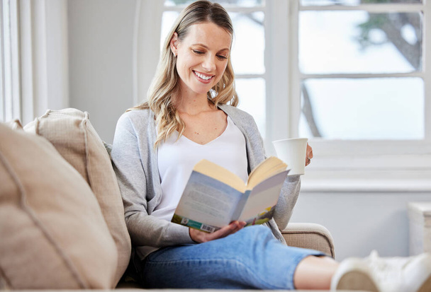 młoda kobieta czytająca książkę podczas relaksu na kanapie w domu. - Zdjęcie, obraz