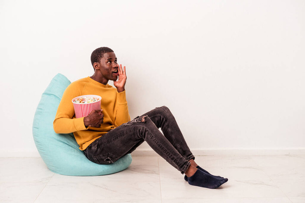 Молодой афроамериканец, сидящий на слоеном попкорне на белом фоне, говорит тайные горячие новости о торможении и смотрит в сторону - Фото, изображение