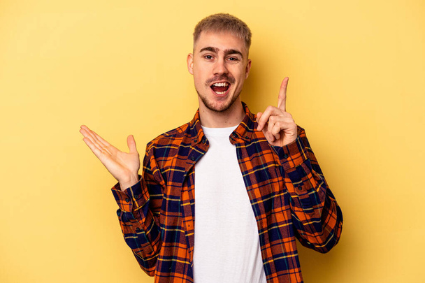 Young caucasian man isolated on yellow background holding and showing a product on hand. - Photo, Image