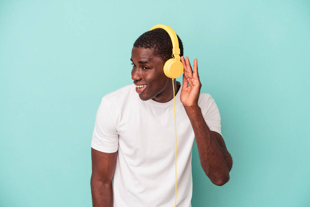 Nuori afroamerikkalainen mies kuuntelemassa sinisellä taustalla eristettyä musiikkia yrittäen kuunnella juoruja. - Valokuva, kuva