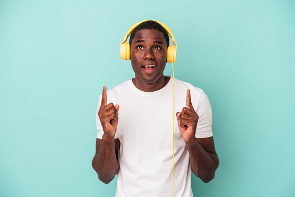 Jeune homme afro-américain écoutant de la musique isolée sur fond bleu pointant à l'envers avec la bouche ouverte. - Photo, image