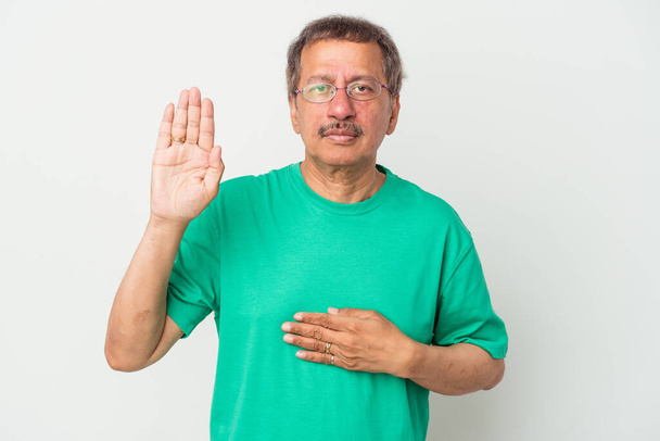 Индийский мужчина средних лет изолирован на белом фоне, давая клятву, положив руку на грудь. - Фото, изображение