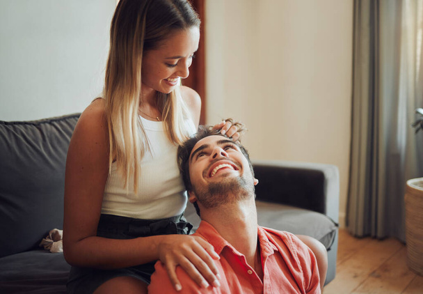 Loving young caucasian couple sitting together at home spending time and happy to be together. Happy young woman sitting on couch while her boyfriend sits between her legs as she plays with his hair - Foto, imagen