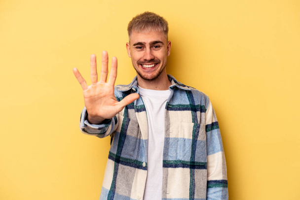 Young caucasian man isolated on yellow background smiling cheerful showing number five with fingers. - Photo, Image