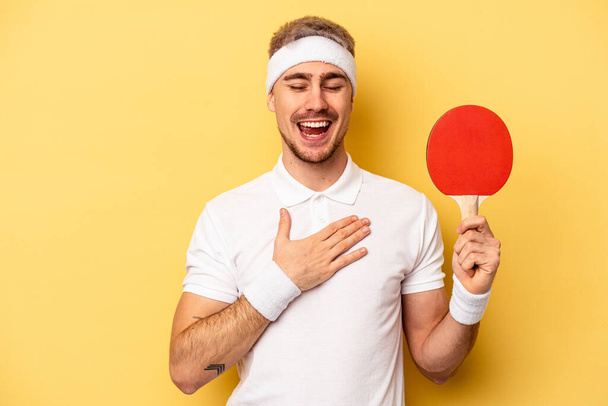 Young caucasian man holding ping pong rackets isolated on yellow background laughs out loudly keeping hand on chest. - Photo, Image