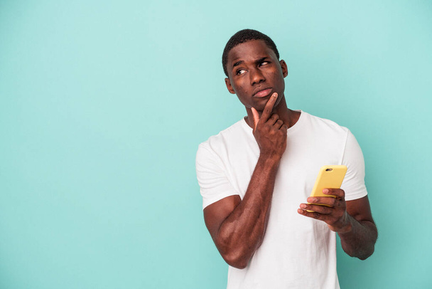 Young African American man holding a mobile phone isolated on blue background looking sideways with doubtful and skeptical expression. - Fotó, kép