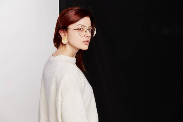 Beautiful redhead model in big eyeglasses posing in black leggings and white sweater on a of half black and white background. Beauty, fashion concept. Business style. - Photo, Image