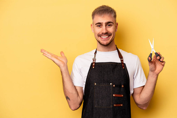 Young caucasian hairdresser man holding scissors isolated on yellow background showing a copy space on a palm and holding another hand on waist. - Foto, afbeelding