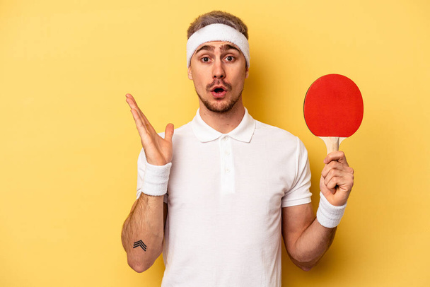 Young caucasian man holding ping pong rackets isolated on yellow background surprised and shocked. - Zdjęcie, obraz