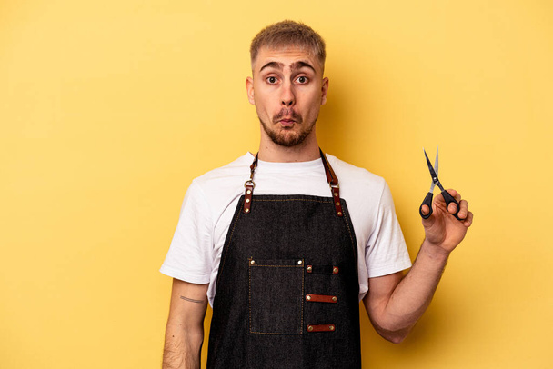 Young caucasian hairdresser man holding scissors isolated on yellow background shrugs shoulders and open eyes confused. - Foto, Bild