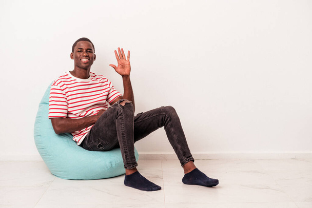 Jonge Afro-Amerikaanse man zit op een trekje geïsoleerd op witte achtergrond glimlachend vrolijk tonen nummer vijf met vingers. - Foto, afbeelding
