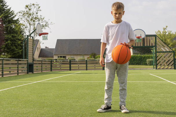 мальчик европейского образца с светлыми волосами в белой футболке и серых брюках стоит на баскетбольной площадке с оранжевым баскетболом. зеленая искусственная трава положена на землю для игры в футбол - Фото, изображение