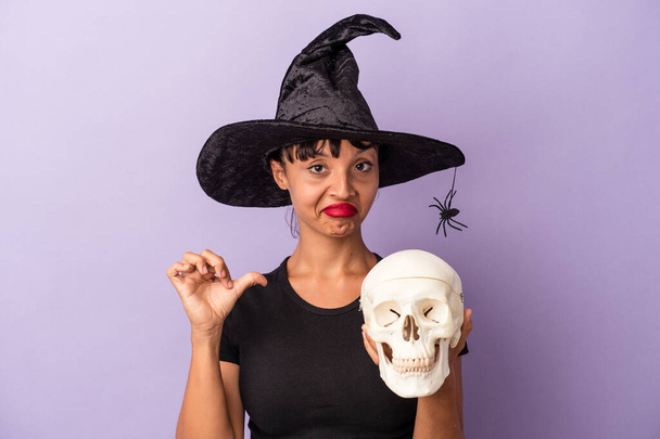 Jeune femme métisse déguisée en sorcière tenant un crâne isolé sur fond violet se sent fière et confiante, exemple à suivre. - Photo, image