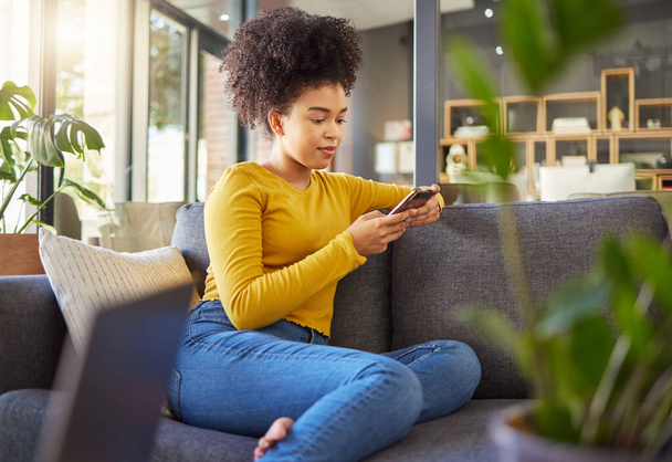 Young mixed race woman typing a message on a phone at home. One hispanic female with a curly afro using social media on a cellphone while relaxing on the couch at home. - Foto, immagini