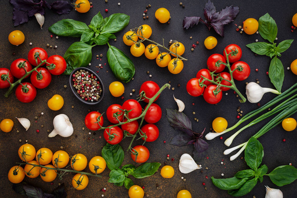 Фон питания, свежие спелые красные и желтые помидоры, специи и листья базилика, чеснок и зеленый лук на темной доске, концепция здоровой пищи, вид сверху - Фото, изображение
