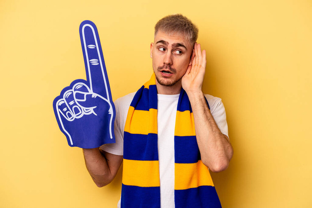 Young sports fan caucasian man isolated on yellow background trying to listening a gossip. - Foto, immagini