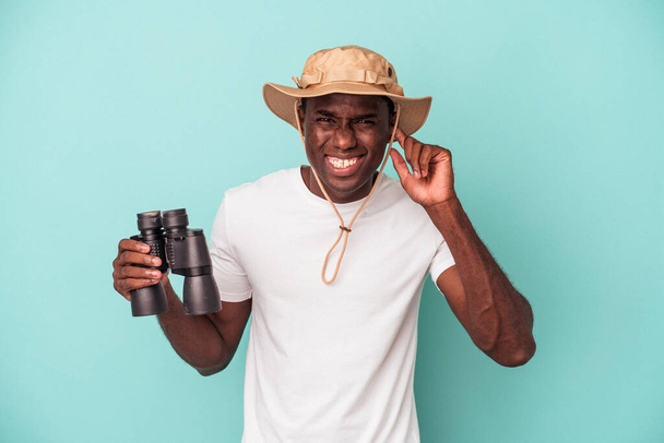 Young African American man holding binoculars isolated on blue background covering ears with hands. - Photo, Image
