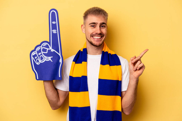 Young sports fan caucasian man isolated on yellow background smiling and pointing aside, showing something at blank space. - Zdjęcie, obraz