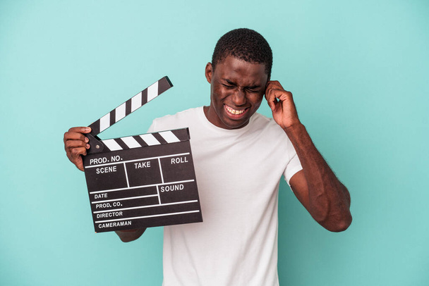 Mladý afroameričan drží clapperboard izolované na modrém pozadí zakrývající uši s rukama. - Fotografie, Obrázek