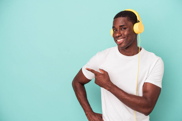 Nuori afrikkalainen amerikkalainen mies kuuntelee sinisellä taustalla eristettyä musiikkia hymyillen ja osoittaen sivuun jotain tyhjää tilaa.. - Valokuva, kuva