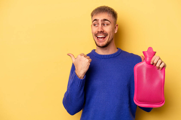 Young caucasian man holding a water bag isolated on yellow background points with thumb finger away, laughing and carefree. - Fotoğraf, Görsel