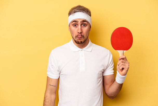 Young caucasian man holding ping pong rackets isolated on yellow background shrugs shoulders and open eyes confused. - Fotó, kép